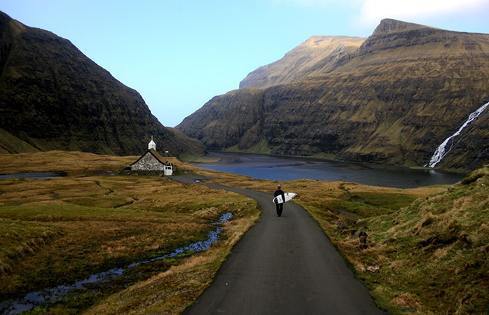 Exact ceea ce vezi, ochii nu te înșeală – un om plimbându-se cu o placă de surf prin satul feroez Saksun © Sergio Villalba & Visit Faroe Islands